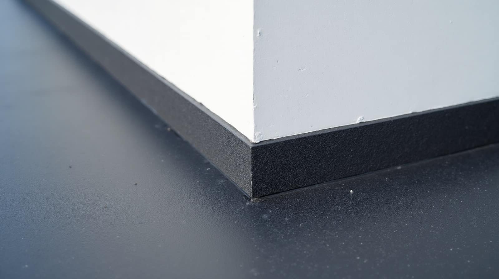 Relevé d'étanchéité où la membrane sombre remonte le pied d'un mur enduit blanc et forme un congé à l'angle